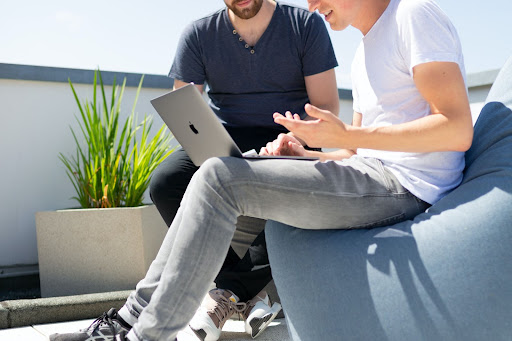 two guys sitting in an open area and discussing while looking at their macbook.