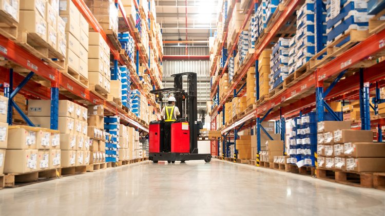 Worker in forklift-truck loading packed goods in huge distribution warehouse with high shelves.